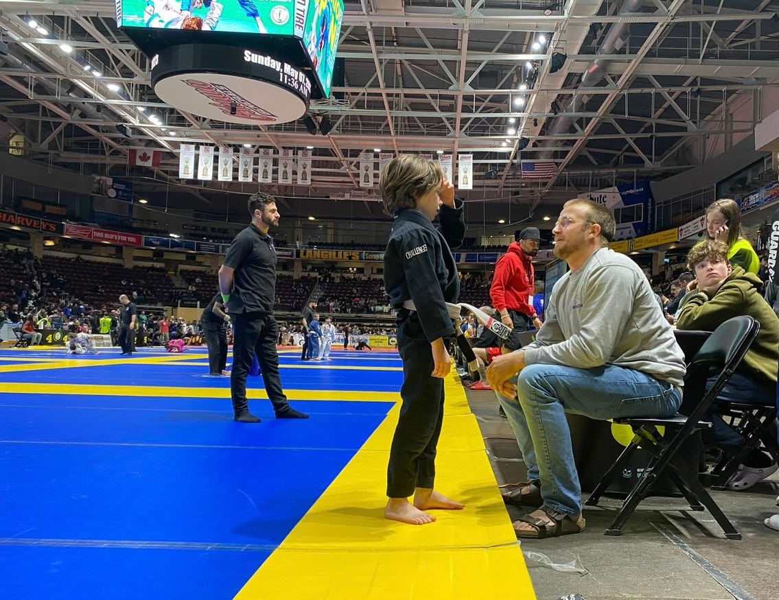 Professor Cody MacCharles giving guidance to a young Brazilian Jiu-Jitsu competitor beside the tournament mats.